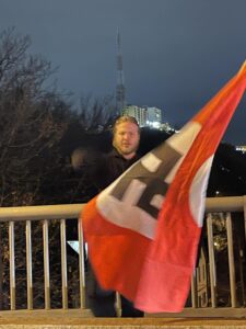 a man with longer blond hair and a beard waves a swastika flag standing in front of a railing