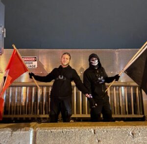 a man with blond hair holds his arms wide with mace and a nazi flag in his hands. a second man, dressed in black, wearing a skull balaclava, holds a black flag